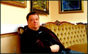 Risto Helin istuu sohvalla, taustalla seinällä tauluja