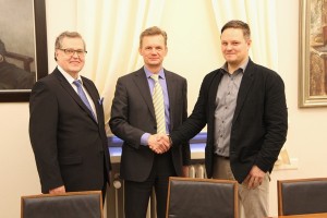 Kolme miestä hymyilee kameralle, kaksi heistä puku päällä