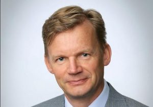 Jari Kuusisto hymyilee kameralle, taustalla Vaasan yliopiston logo